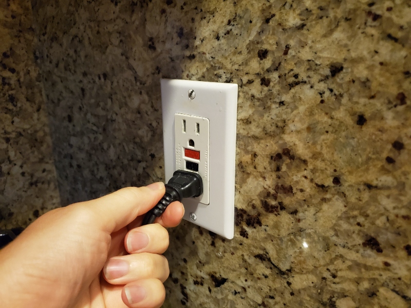 hand plugging in black electrical cord to a white GFCI outlet on a speckled tile wall